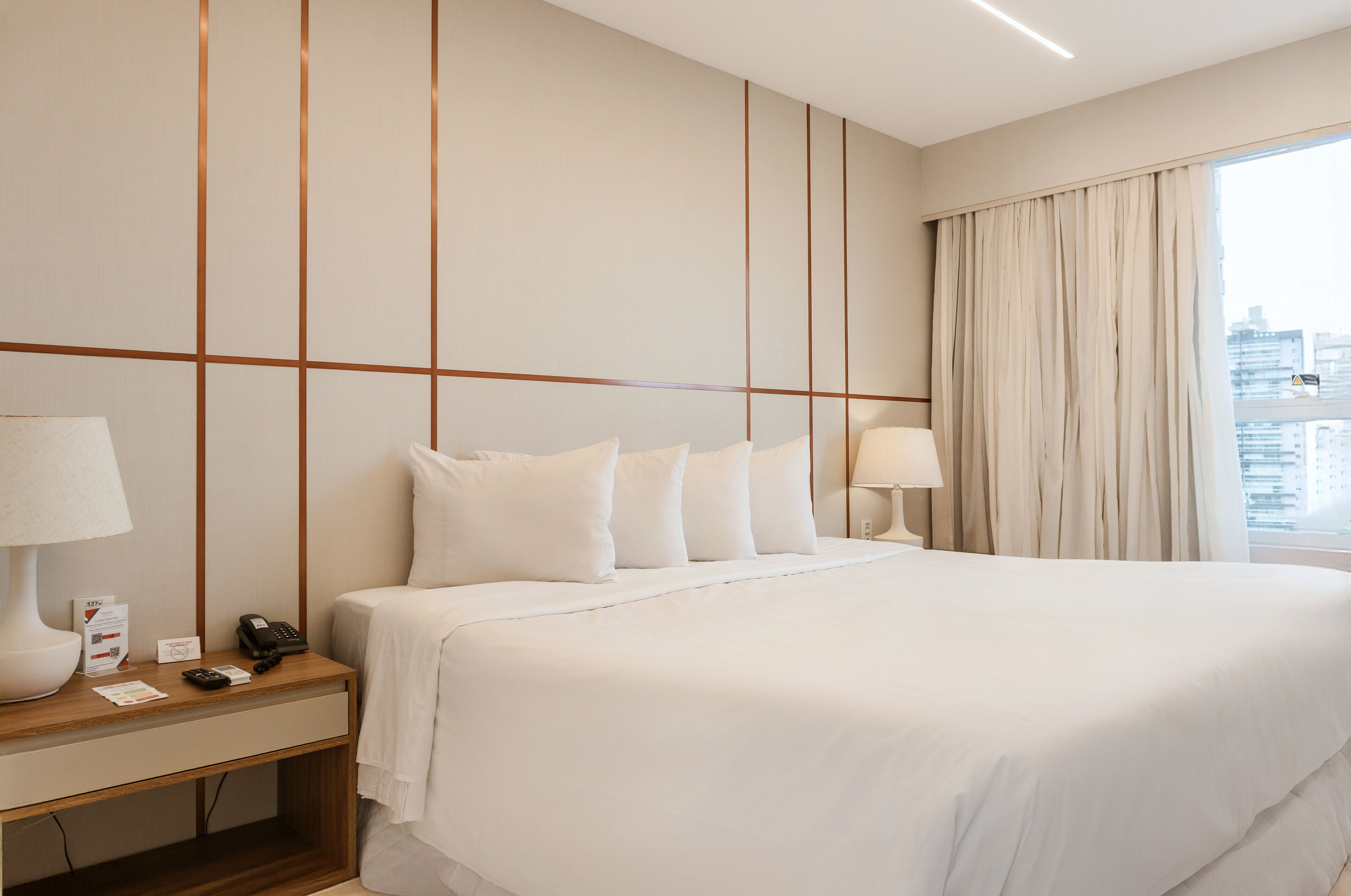 A hotel room with a large bed, a bedside table with a lamp, a phone, and a sign.