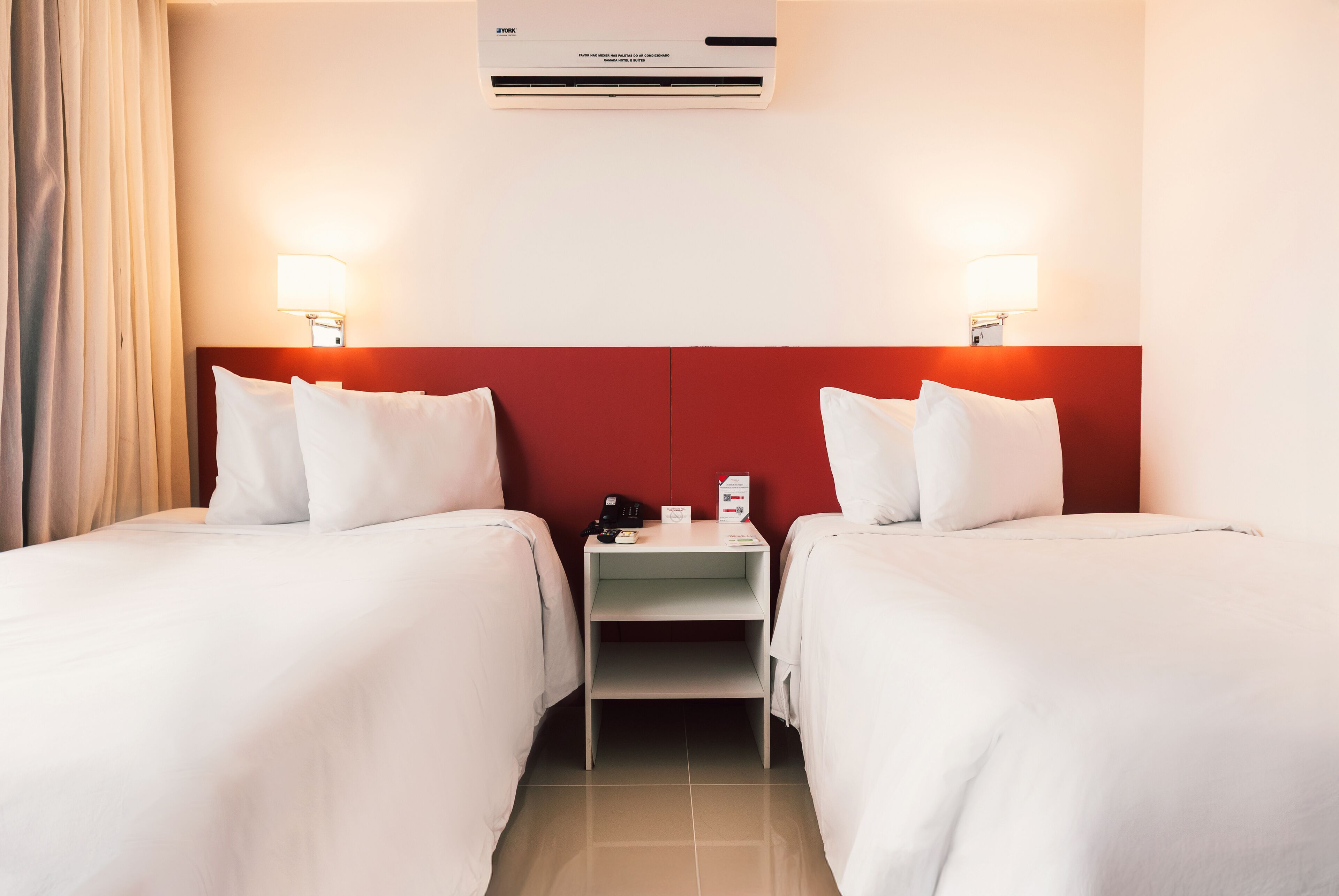 Habitación de hotel con dos camas, un cabecero rojo, una mesita de noche con lámpara y un equipo de aire acondicionado.