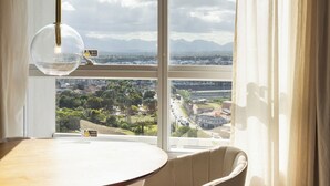 Un comedor con una mesa redonda y dos sillas, ventanas amplias con cortinas, y vistas a un paisaje urbano y montañas.