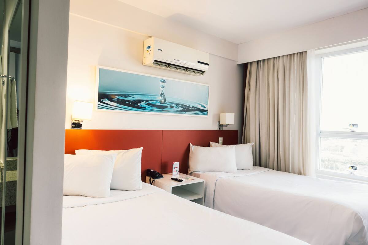Habitación de hotel con dos camas, un ventanal grande con cortinas, un equipo de aire acondicionado, un cuadro de agua y una mesita de noche con teléfono y control remoto.