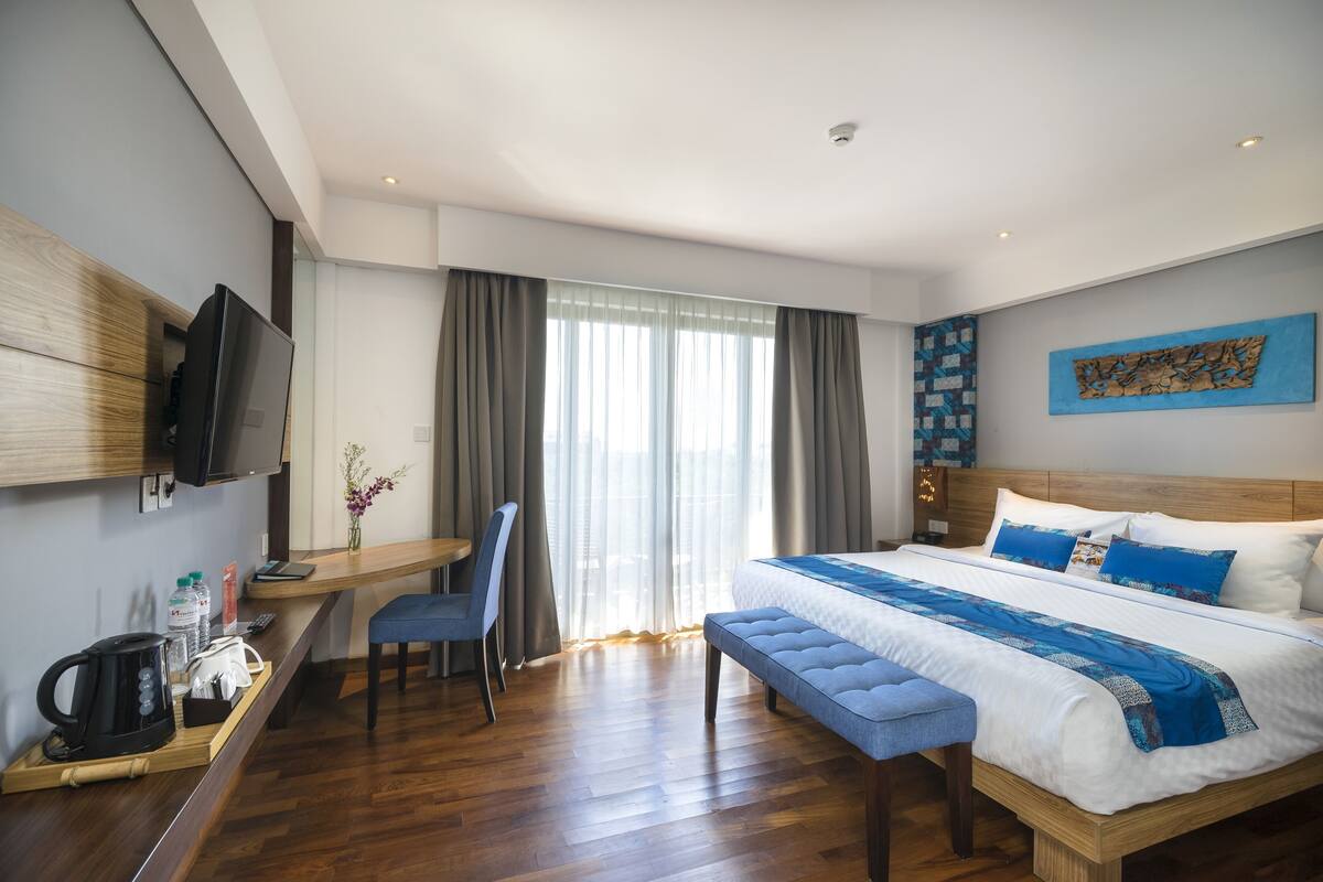 A modern hotel room with a large bed, a desk with a chair, a television, and a window with curtains.