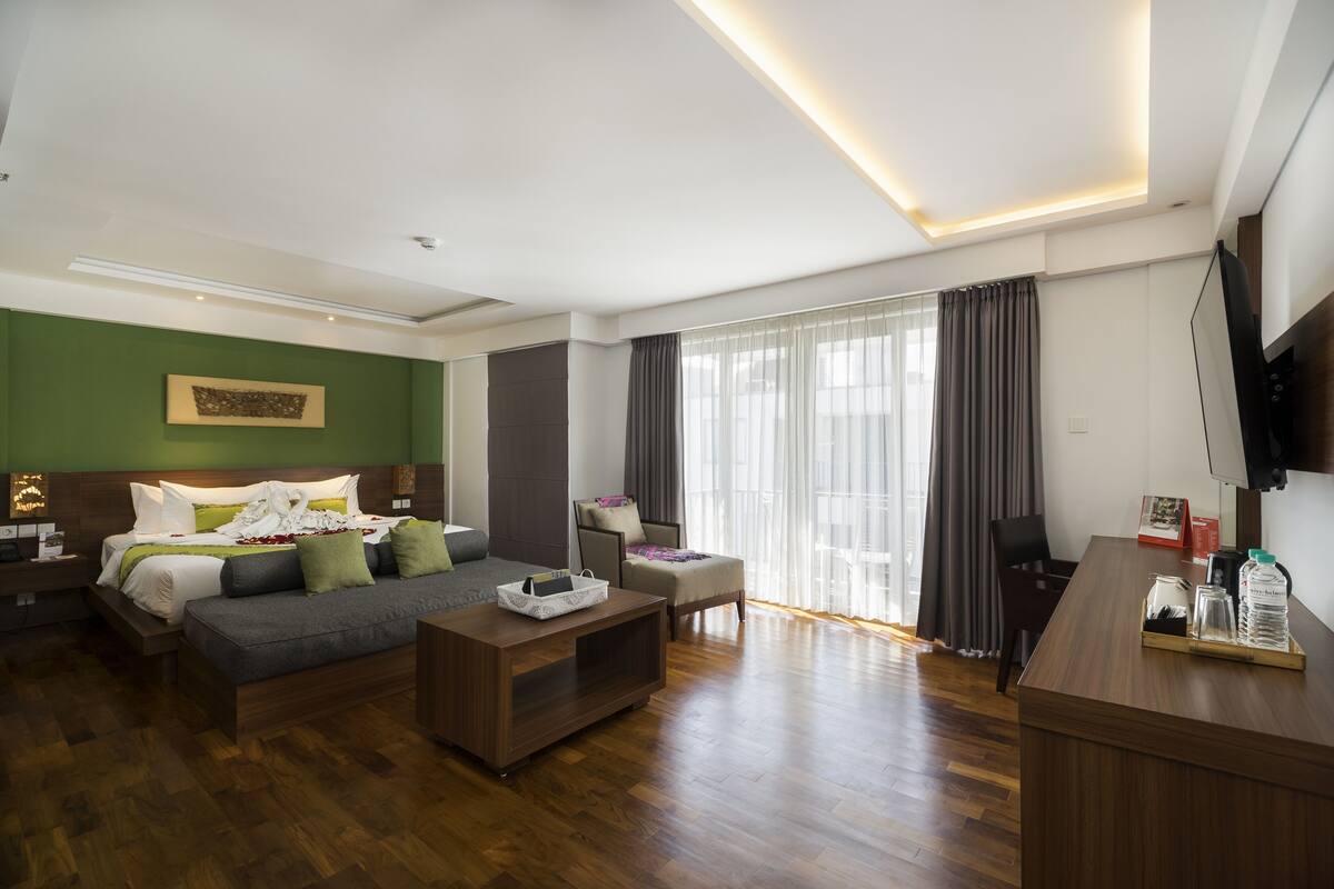 A modern hotel room with a wooden floor, a bed with green and white bedding, a gray sofa, a wooden coffee table, a desk with a chair, and a large window with curtains.