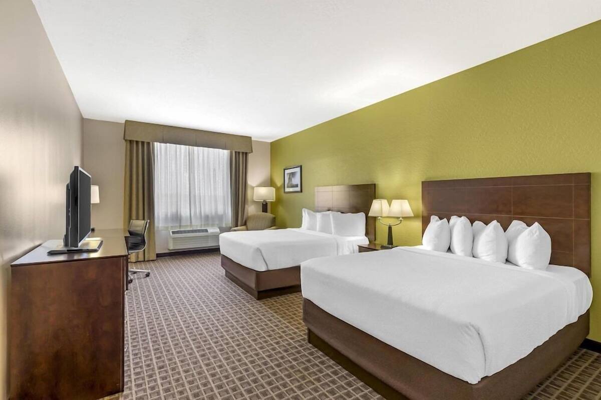 Habitación de hotel con dos camas, un escritorio con televisor, una silla, una ventana con cortinas y un cuadro en la pared.