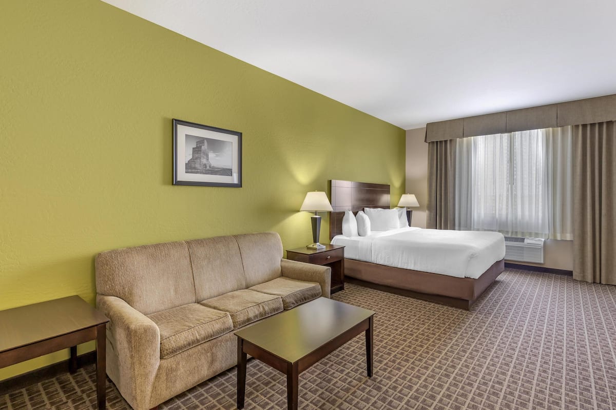 Una habitación de hotel con pared verde, una cama con ropa blanca, un sofá color beige, una mesita pequeña y una lámpara.