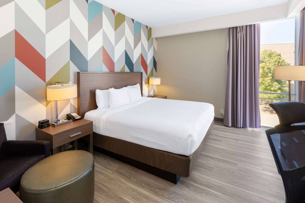 A hotel room with a large bed, a desk, a chair, a lamp, and a wall with a geometric pattern.