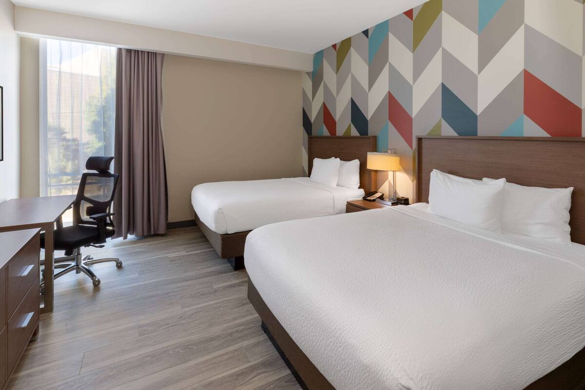 A hotel room with two beds, a desk, a chair, and a window with curtains.