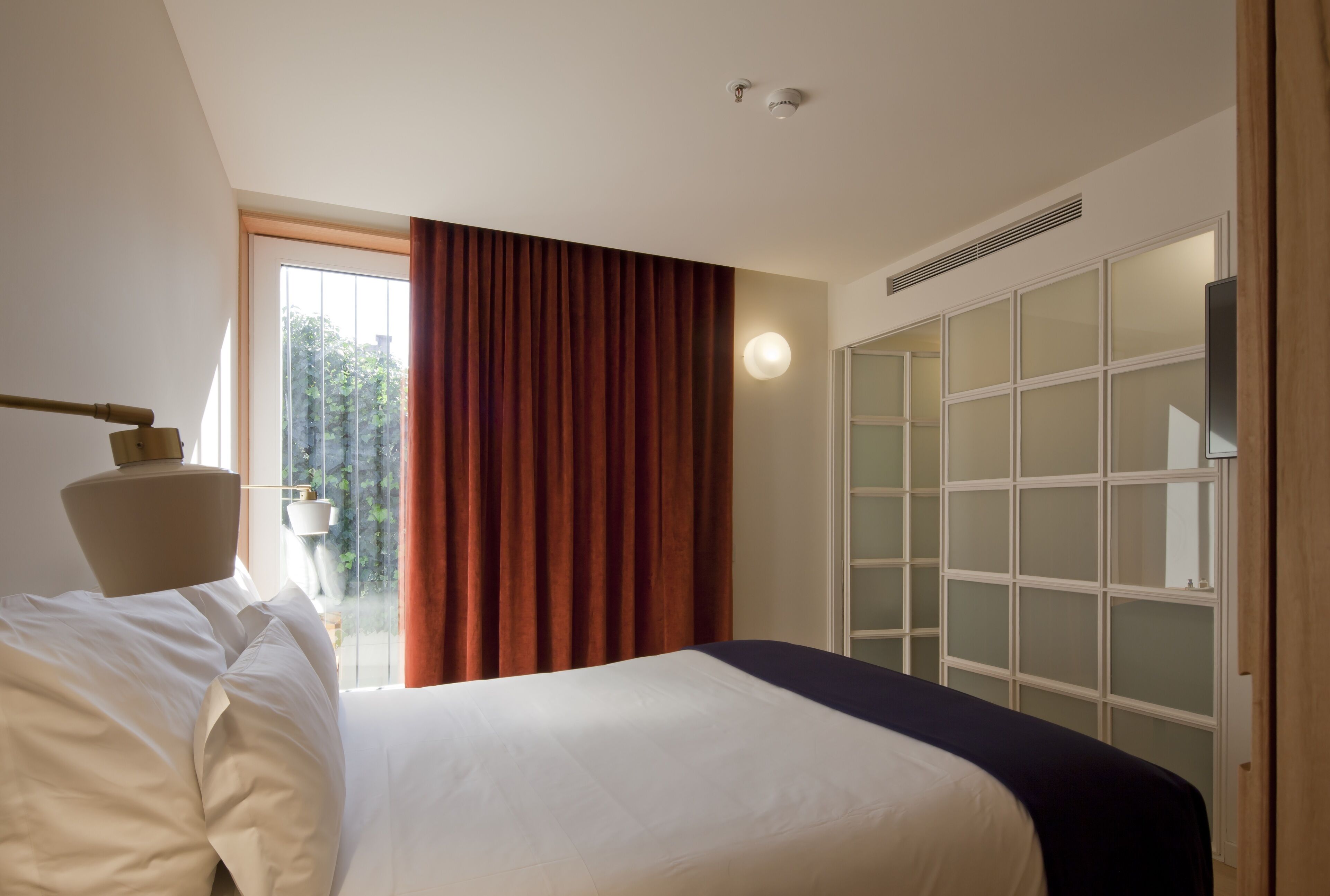 A hotel room with a bed, a lamp, a window with curtains, and a wardrobe with frosted glass doors.