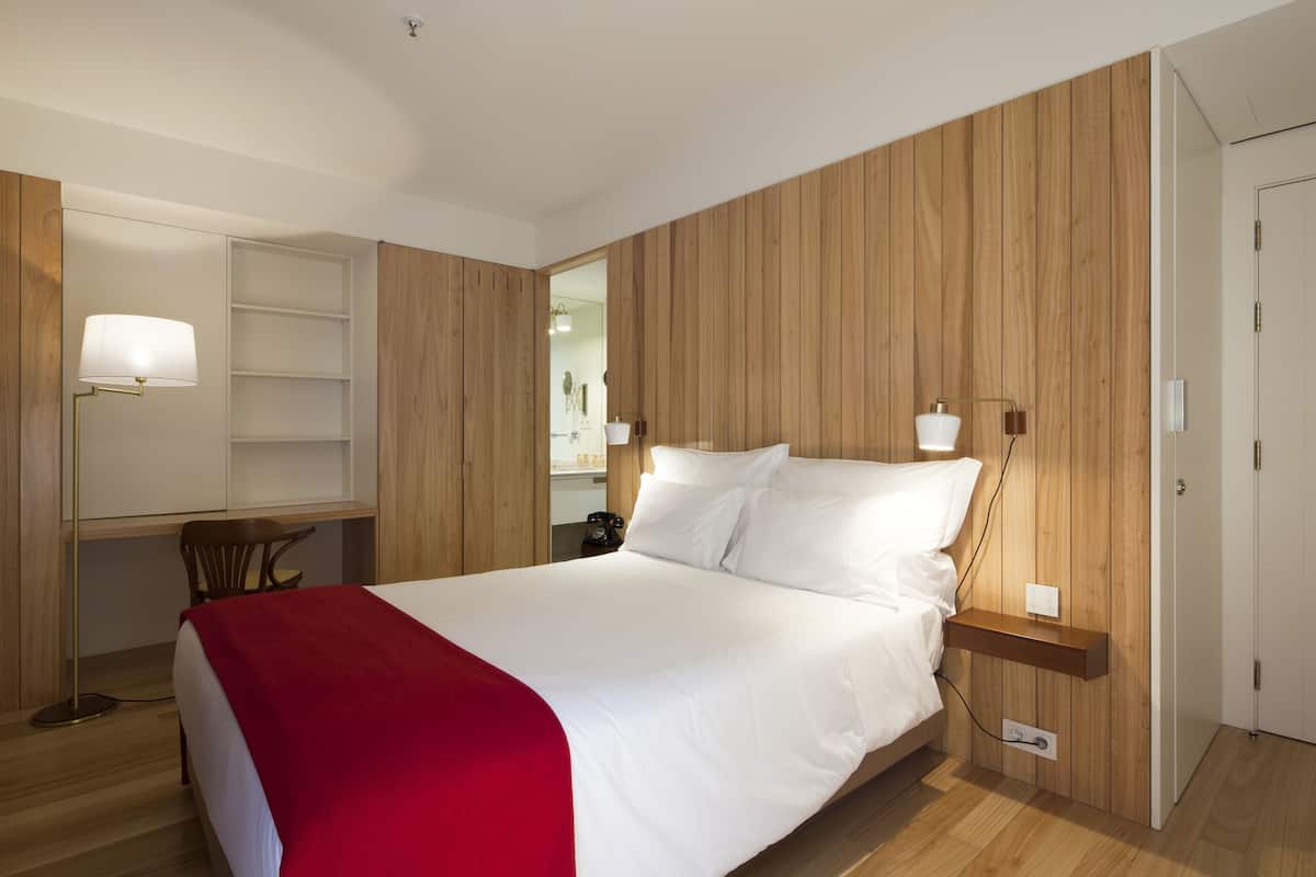 A modern bedroom with a large bed, wooden panel walls, a desk with a chair, and bedside lamps.