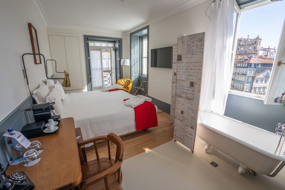 A hotel room with a bed, a desk with a computer, a chair, a bathtub, and a window with a city view.