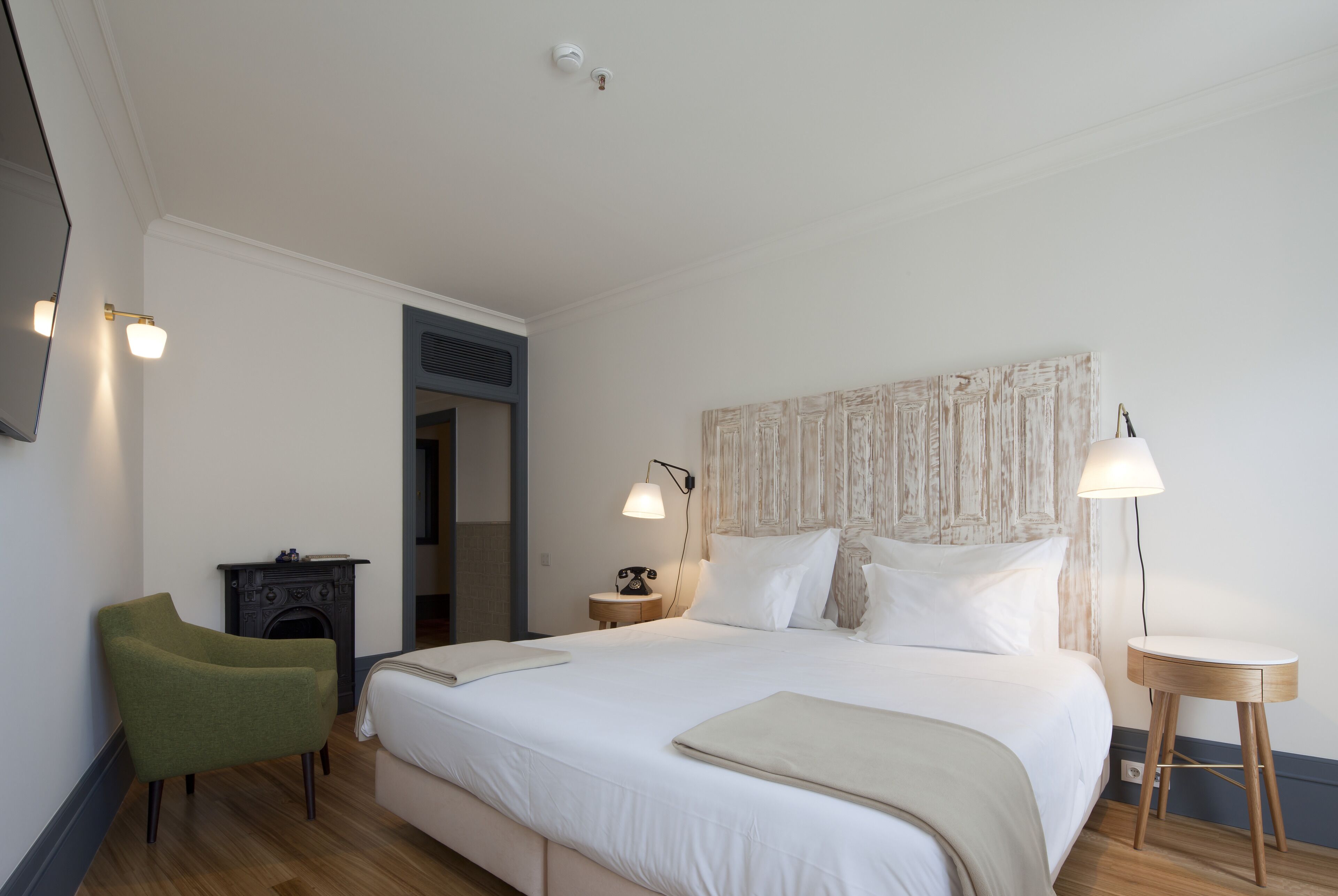 A hotel room with a large bed, a green armchair, a small table, and a fireplace.