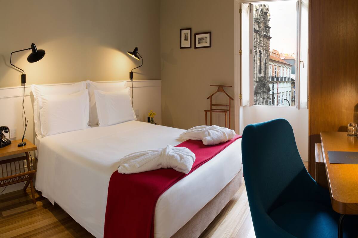 A hotel room with a bed, a desk, a chair, a desk lamp, a telephone, and a window with a view of buildings.