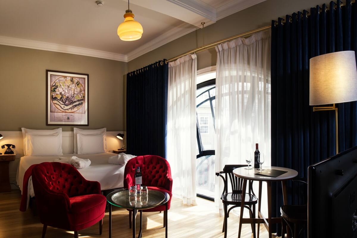 A hotel room with a bed, a red armchair, a small round table with glasses, a lamp, and a framed map on the wall.