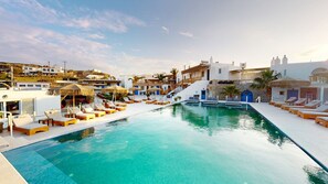 Un área de piscina con sillones de descanso y una piscina azul cristalina, rodeada de edificios blancos con detalles azules y una escalera que lleva a un nivel superior.