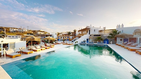 A pool area with lounge chairs and a clear blue pool, surrounded by white buildings with blue accents and a staircase leading to an upper level.