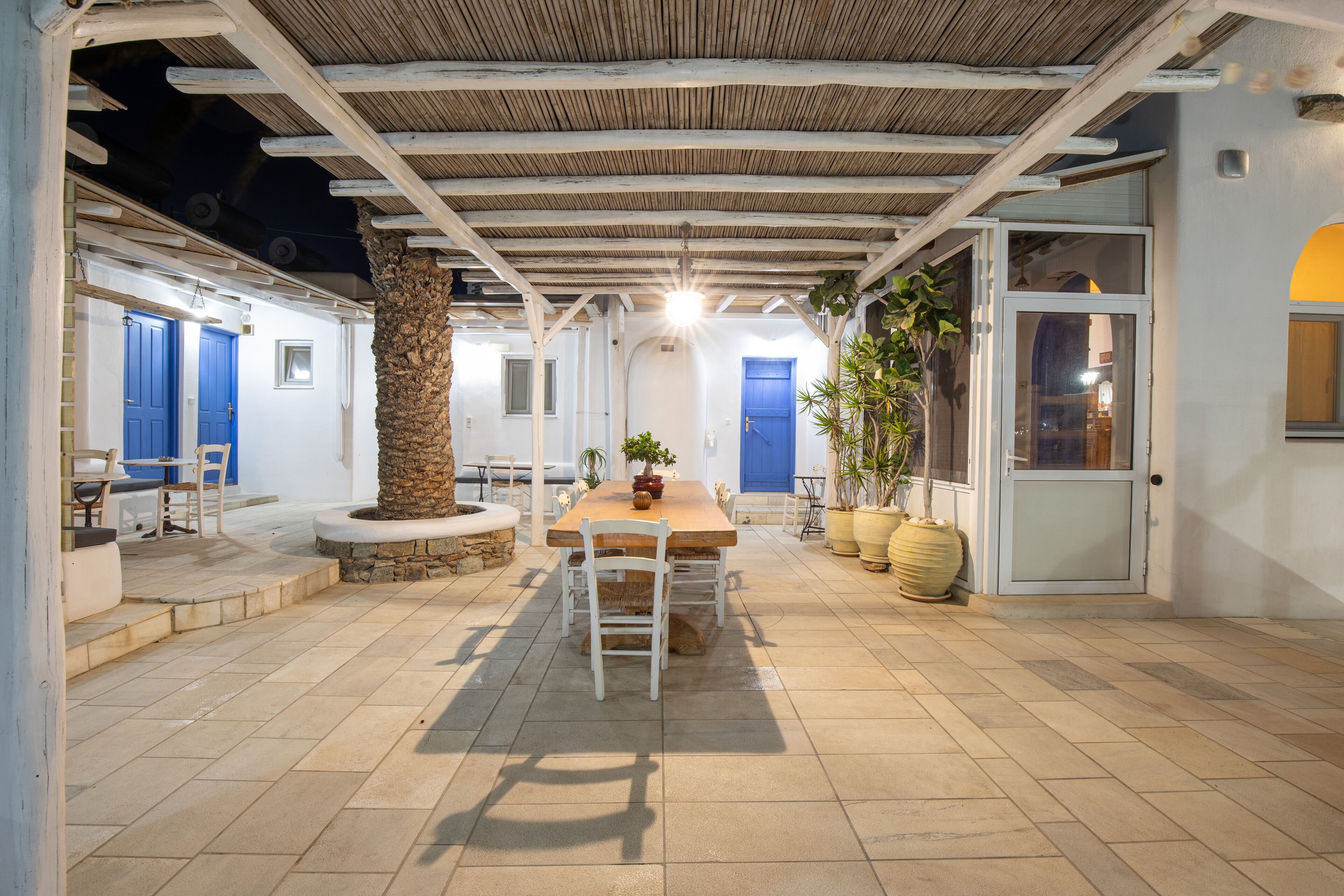 An outdoor dining area with a wooden table and chairs, surrounded by potted plants and a stone pillar.