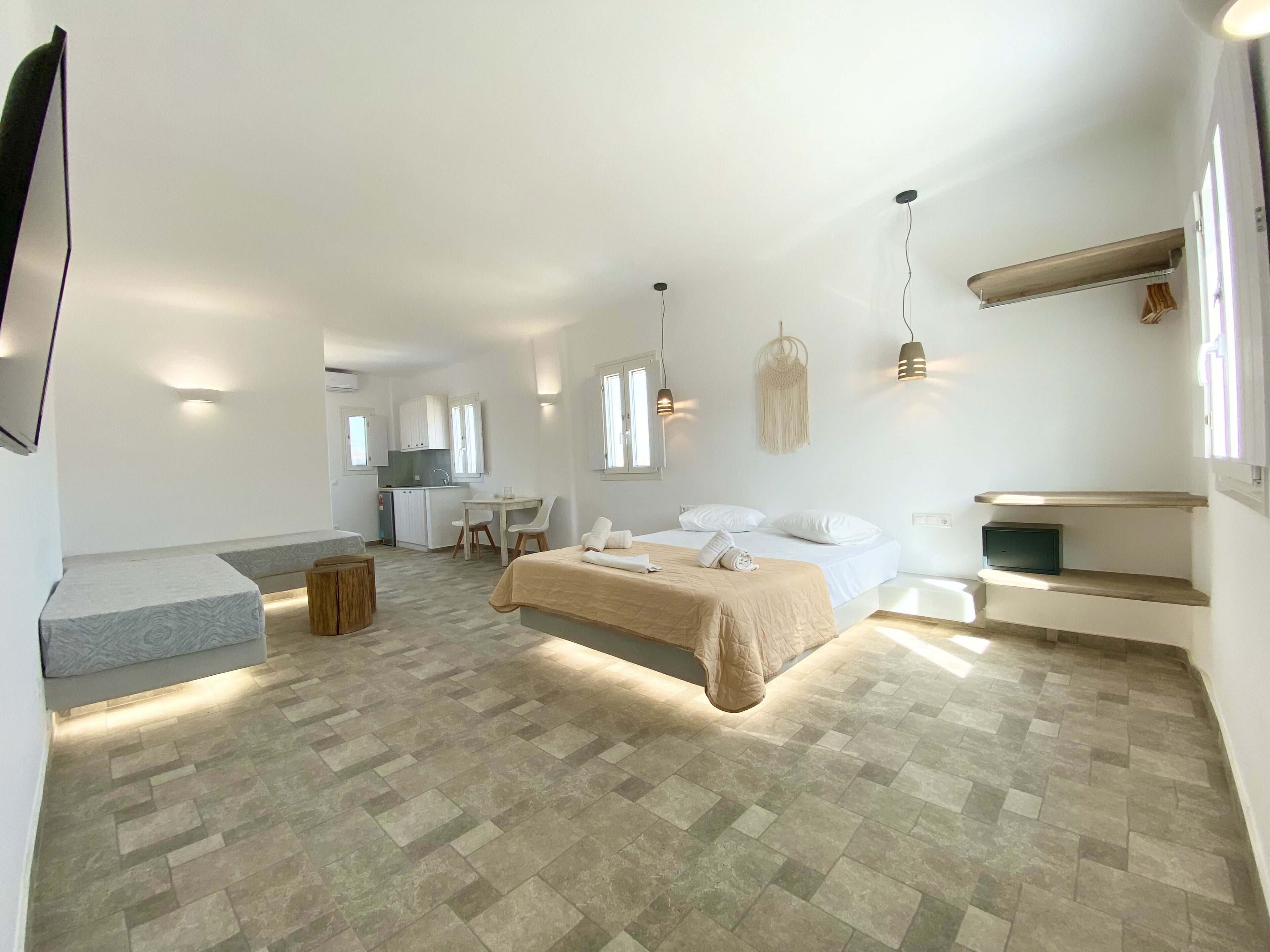 Quarto moderno e bem iluminado, com uma cama grande, uma escrivaninha pequena e uma cozinha compacta ao fundo.