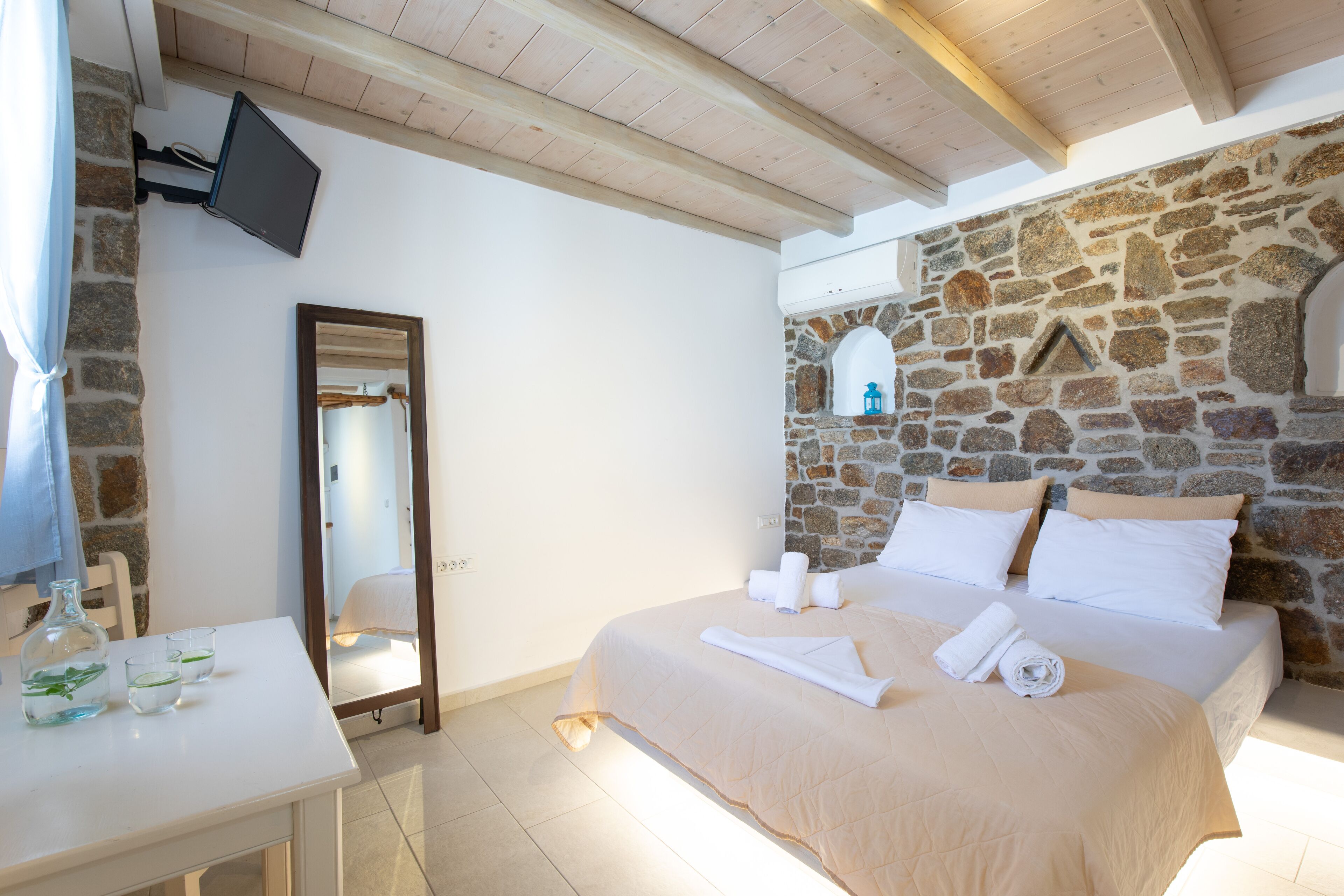 A bedroom with a stone wall, a bed with white linens, a flat-screen TV mounted on the wall, and a small table with a bottle and glasses.
