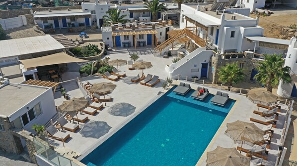 Un área de piscina en la azotea con sillones y sombrillas, rodeada de edificios blancos con puertas y ventanas azules.