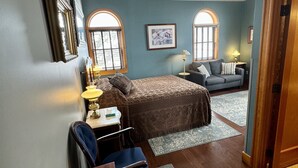 Un dormitorio acogedor con cama, escritorio, silla y sofá. Hay dos ventanas con arcos, una lámpara y un cuadro en la pared.