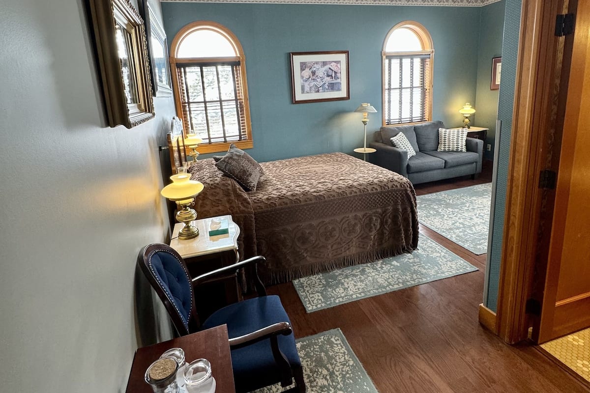 A cozy bedroom with a bed, a desk, a chair, and a sofa. There are two windows with arched tops, a lamp, and a framed picture on the wall.