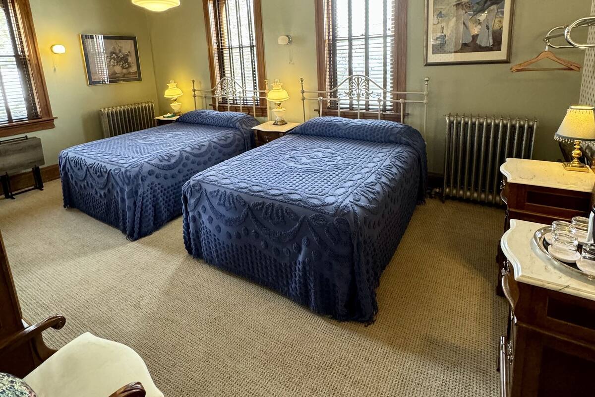 A hotel room with two beds, a desk, a chair, a radiator, and a window with a view.