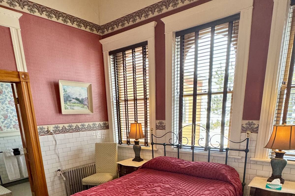 A bedroom with a large bed, two bedside tables with lamps, a radiator, and a window with blinds.