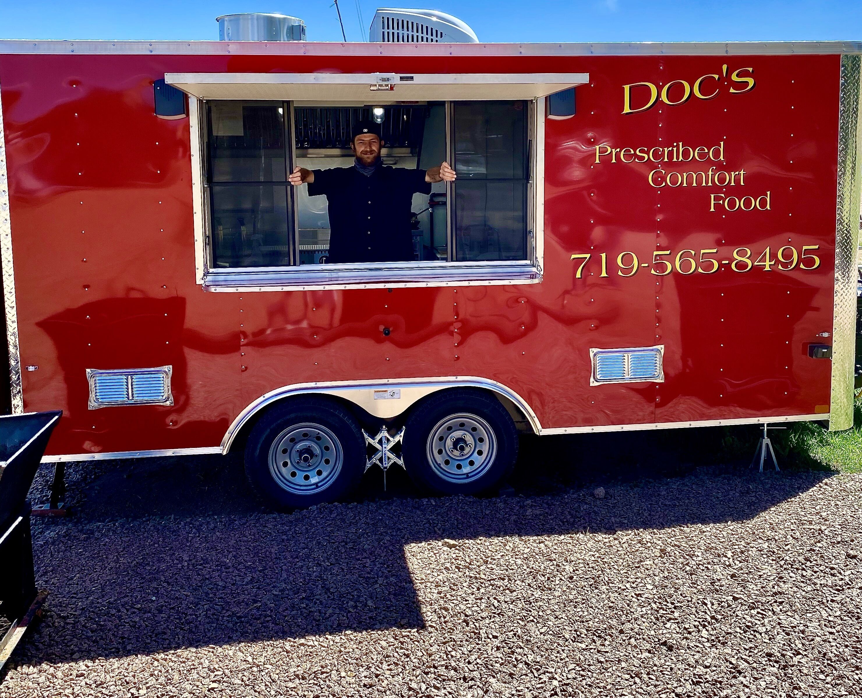 Een rode foodtruck met een raam, met de tekst 'Doc's Prescribed Comfort Food' en het telefoonnummer 719-565-8495.