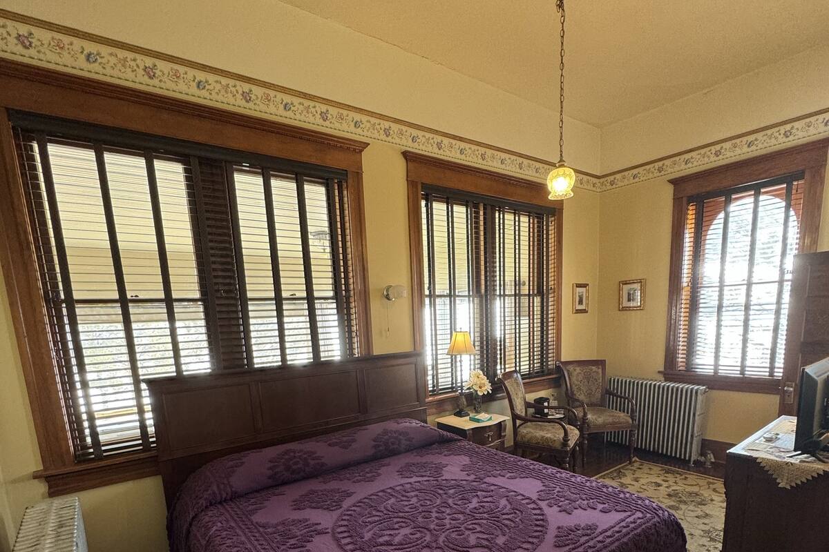 A room with a large bed, wooden flooring, and a radiator. There are windows with blinds, a desk, and a chair.