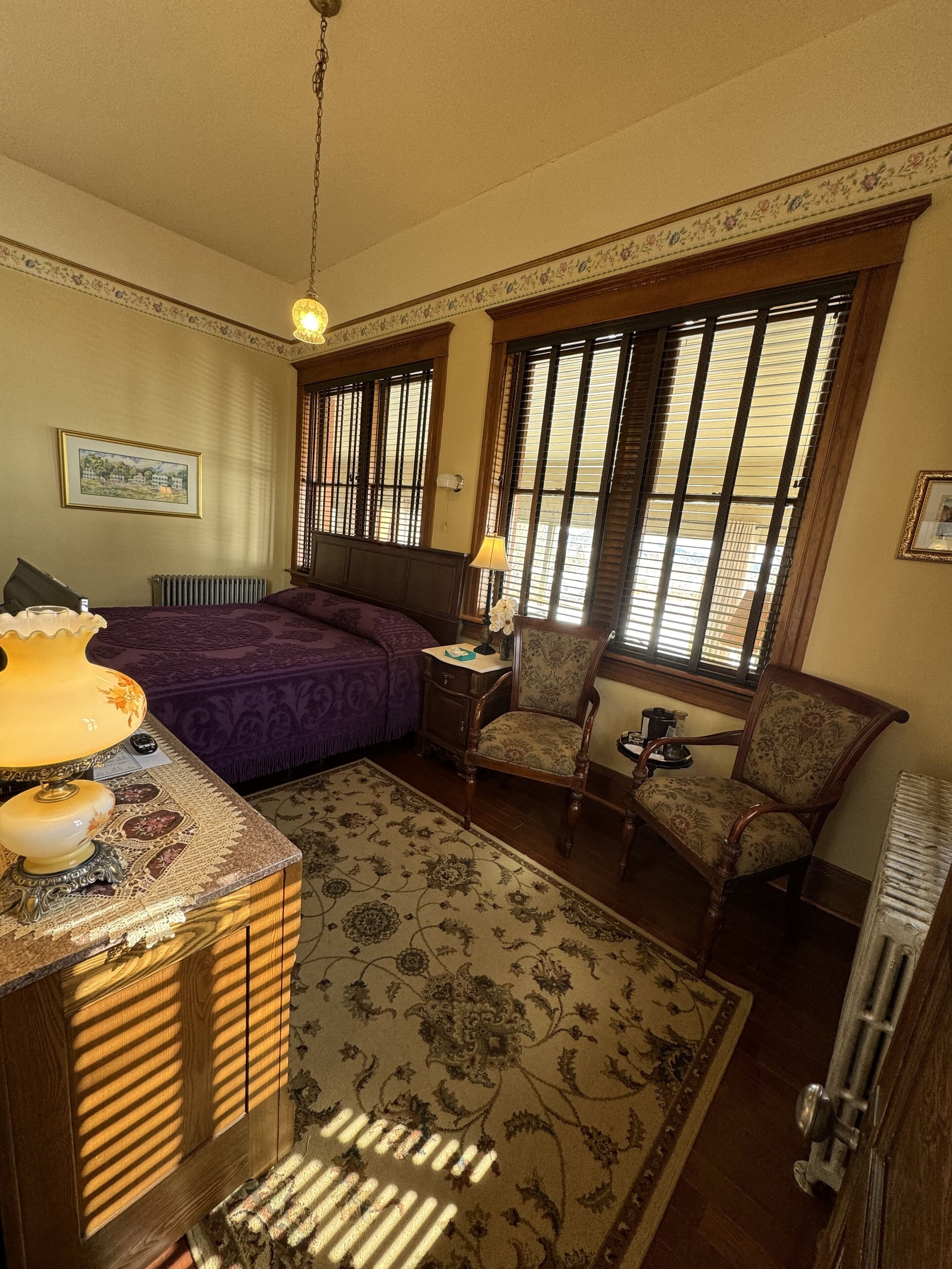 A bedroom with a bed, a chair, a nightstand, a lamp, a radiator, and a window with blinds.
