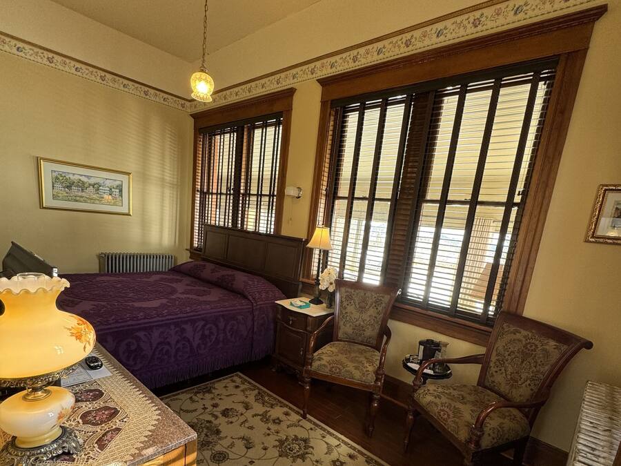 A bedroom with a bed, a chair, a nightstand, a lamp, a radiator, and a window with blinds.