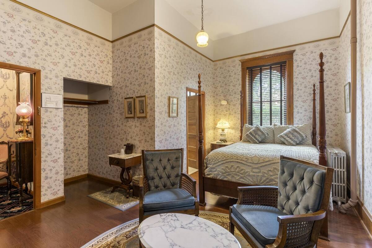 A bedroom with a four-poster bed, two upholstered chairs, a small table, and a radiator.