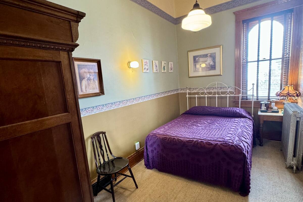 A bedroom with a bed, a chair, a dresser, a radiator, and a window with curtains.