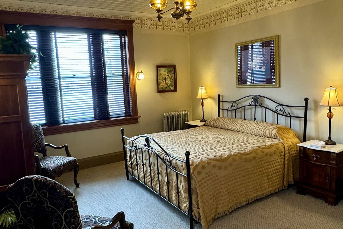 A bedroom with a four-poster bed, a chandelier, a radiator, and a window with blinds.