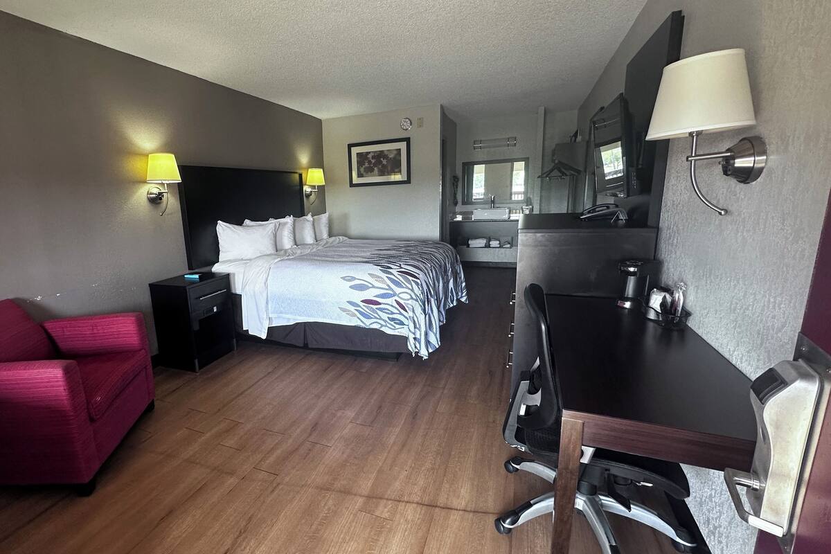 Habitación de hotel con cama, escritorio, silla, lámpara de escritorio, mesita de noche, televisor montado en la pared y un marco de fotos.