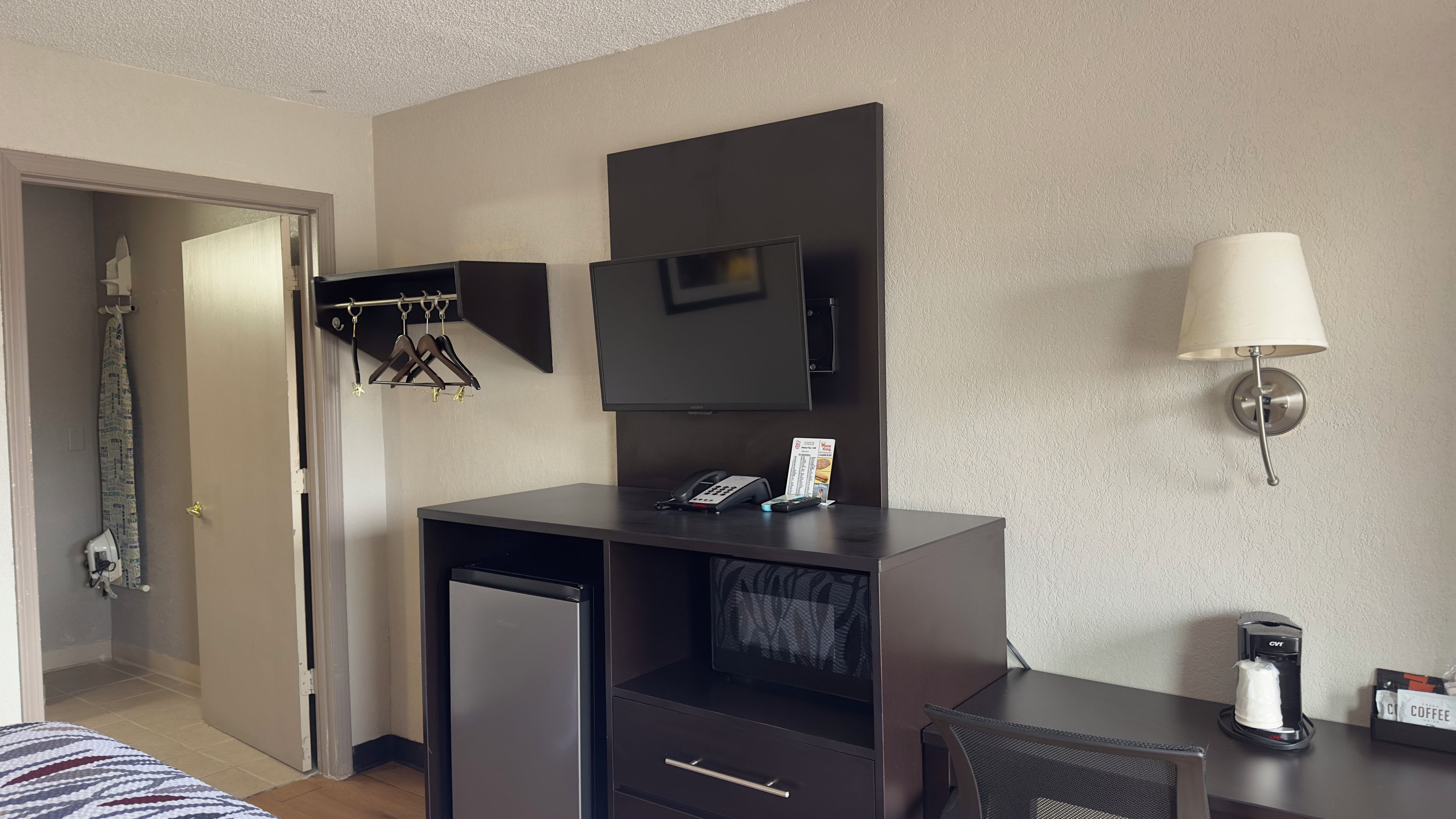 A hotel room with a desk, a TV, a lamp, a coffee maker, and a hanging coat rack.