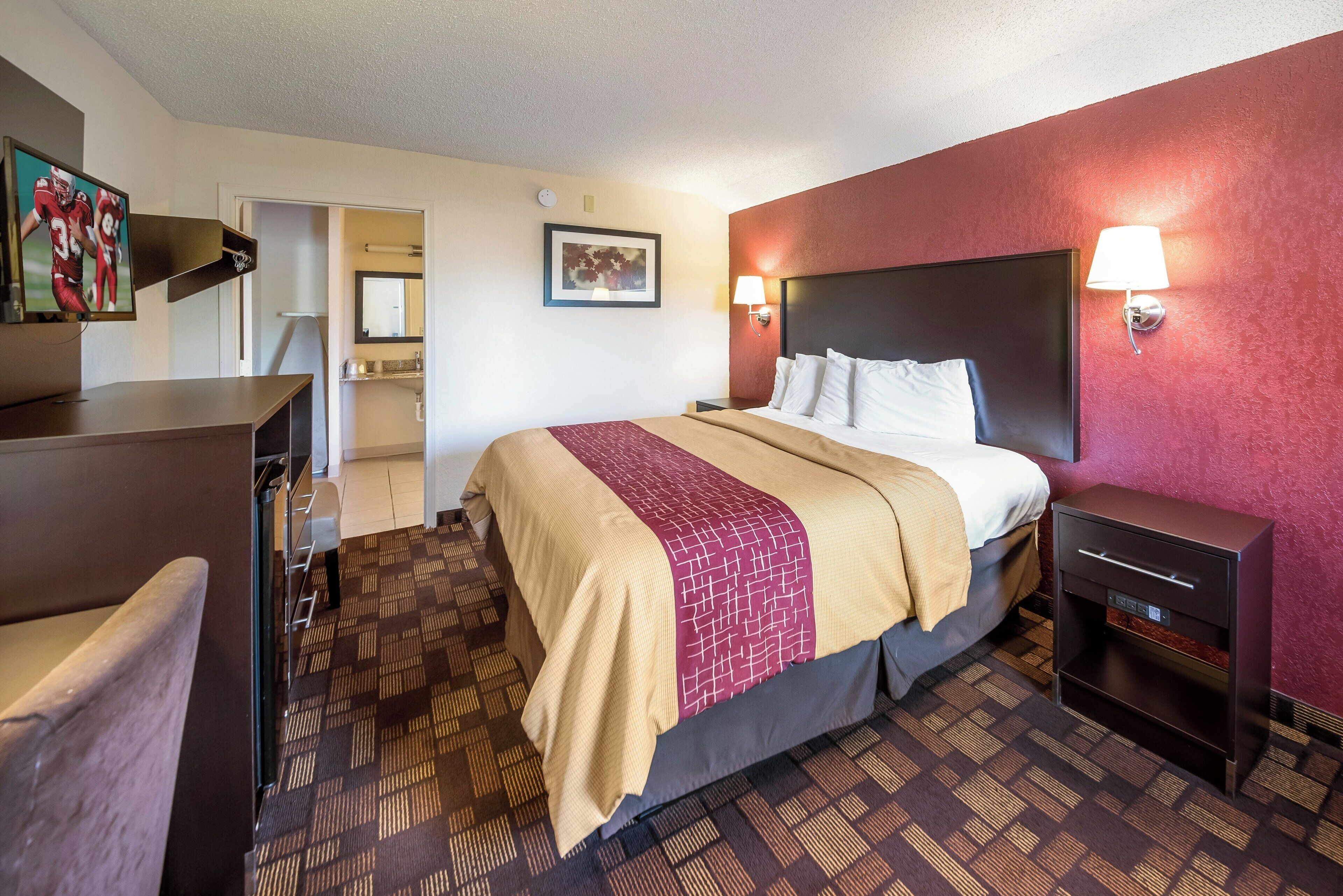 A hotel room with a bed, bedside table, lamp, and a desk with a computer.