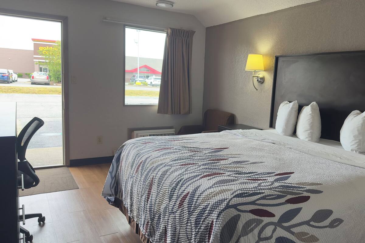 Habitación de hotel con una cama grande, un escritorio, una silla, una ventana con vista a un estacionamiento y una lámpara fijada a la pared.
