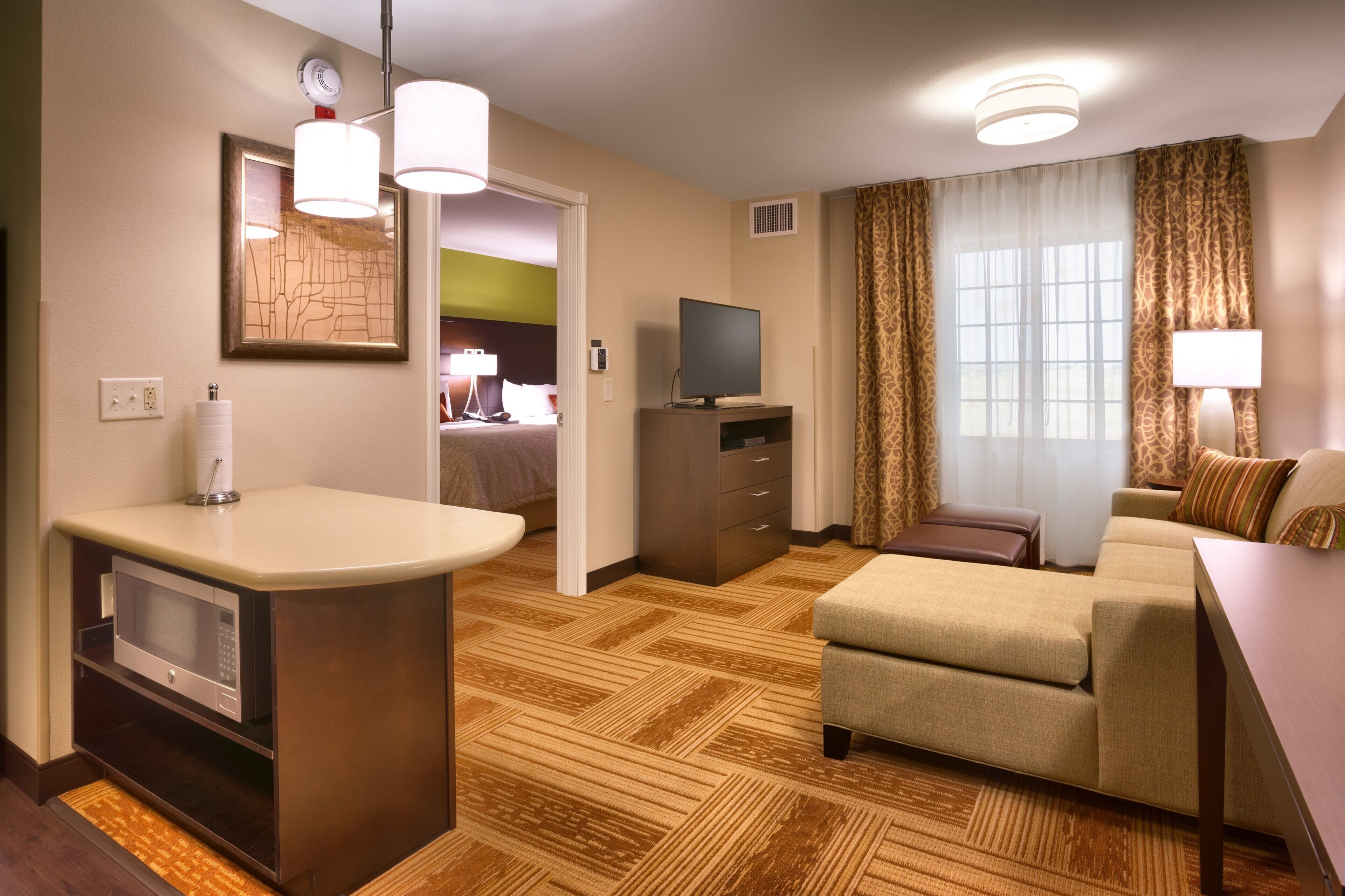 Habitación de hotel con kitchenette, televisor, sofá y ventana con cortinas.