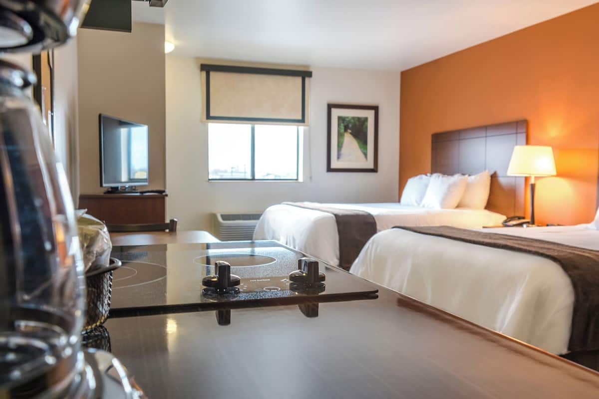 A hotel room with two beds, a TV, a window with blinds, a framed picture on the wall, and a bedside lamp.