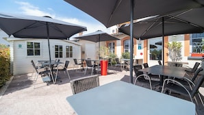 An outdoor patio area with tables and chairs under large umbrellas, a white building with windows, and a small red trash bin. - Hotel du Lauragais (Villefranche-de-Lauragais)