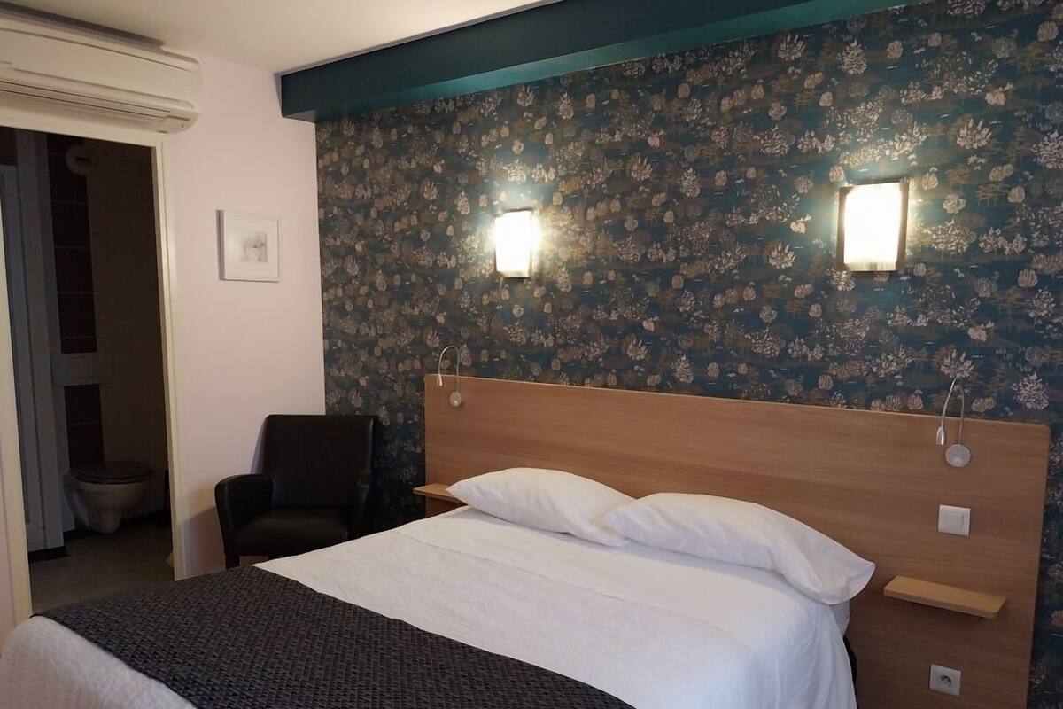 Habitación de hotel con cama, silla, mesita de noche, lámpara de pared y unidad de aire acondicionado.
