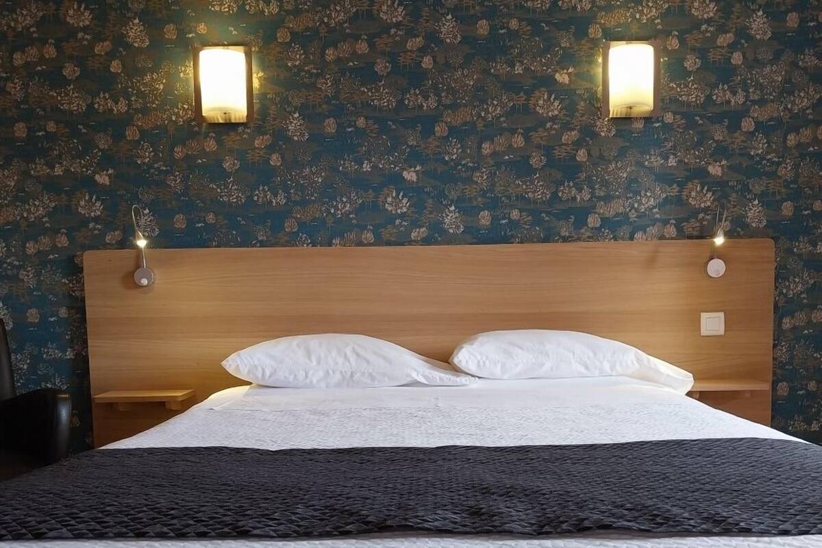 Una cama bien hecha con sábanas blancas y una manta oscura, un cabecero de madera y un papel tapiz floral con dos luces fijadas a la pared.
