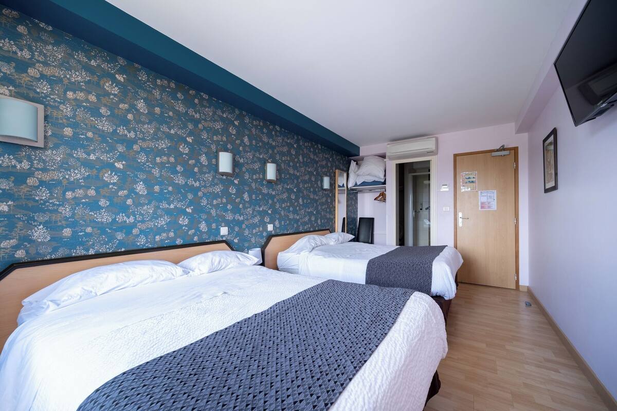 Habitación de hotel con dos camas, un papel tapiz floral azul, una puerta de madera y un televisor en la pared.
