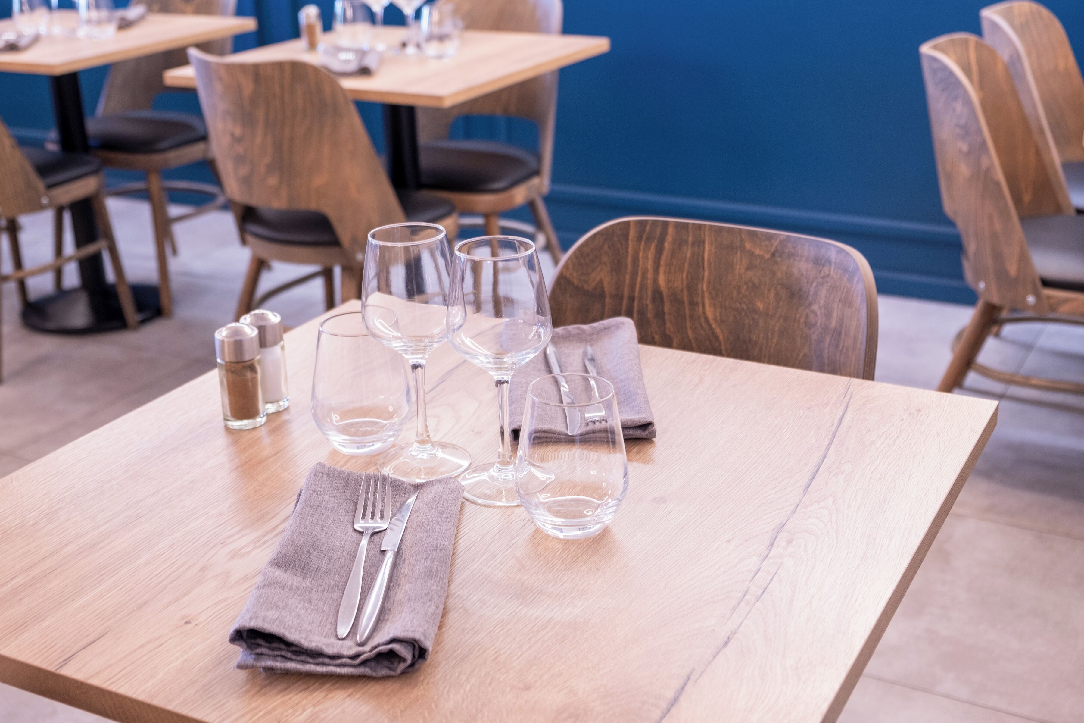 Mesa posta com talheres e copos de vinho, com cadeiras e mesas ao fundo.