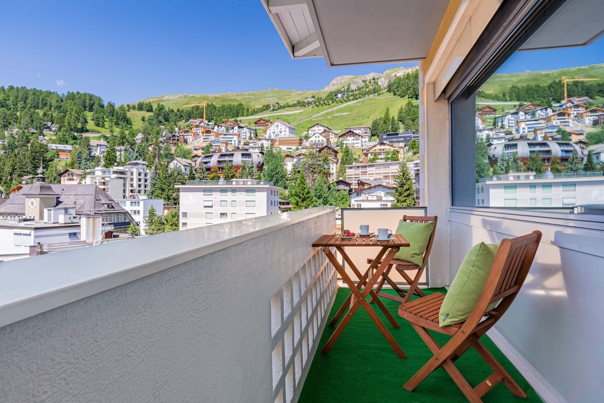 A balcony with a view of a hillside town, wooden chairs, and a small table.