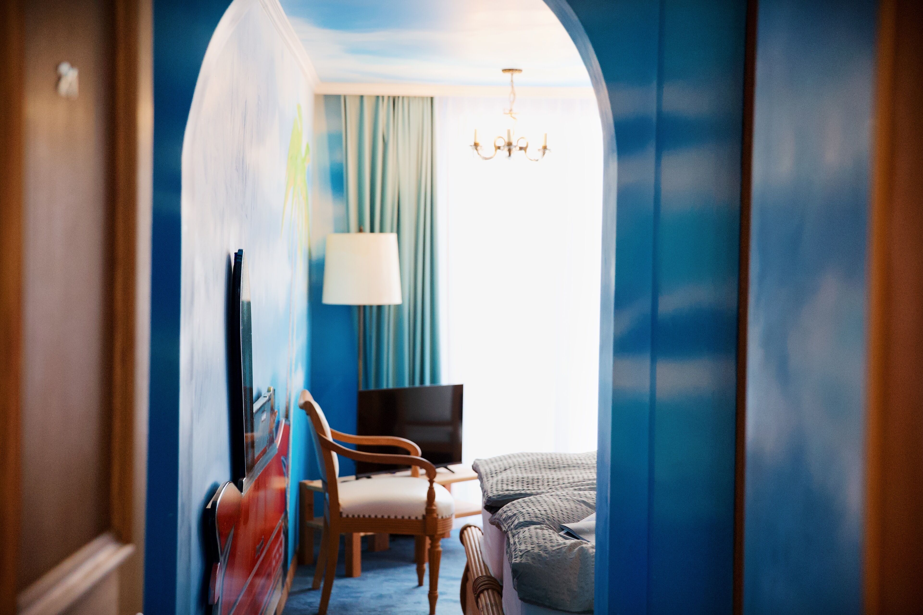 Habitación de hotel con pared azul, una lámpara de araña, cama, silla, escritorio y televisor.