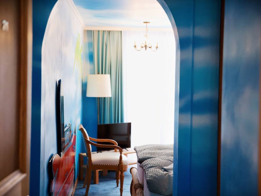 Habitación de hotel con pared azul, una lámpara de araña, cama, silla, escritorio y televisor.