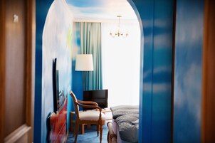 Habitación de hotel con pared azul, una lámpara de araña, cama, silla, escritorio y televisor.