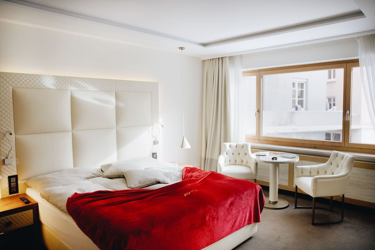 A hotel room with a large bed, a red blanket, a white chair, a small round table, and a window with curtains.