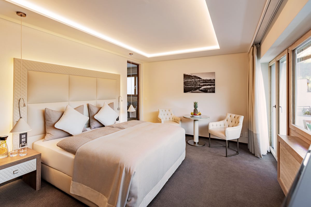 A modern hotel room with a large bed, bedside tables, a small round table, a chair, and a window with curtains.